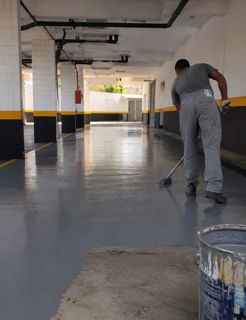 Trabalhador aplicando impermeabilização epóxi cinza em piso de garagem predial, movimentando rodo em corredor amplo com colunas brancas e faixa amarela e preta de sinalização, balde de tinta em primeiro plano, iluminação natural difusa, fotografia realista de obra predial