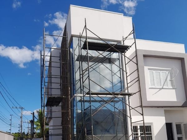 Fachada de sobrado em construção com andaimes metálicos e fechamento em vidro, estrutura branca moderna, céu azul com nuvens, fotografia realista de obra em fachada