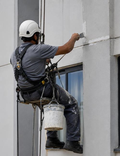 Profissional realizando pintura predial suspenso por cadeira de rapel, capacete de segurança, aplicando tinta na fachada de prédio, cinto e cordas de segurança, fotografia realista de pintura predial e comercial