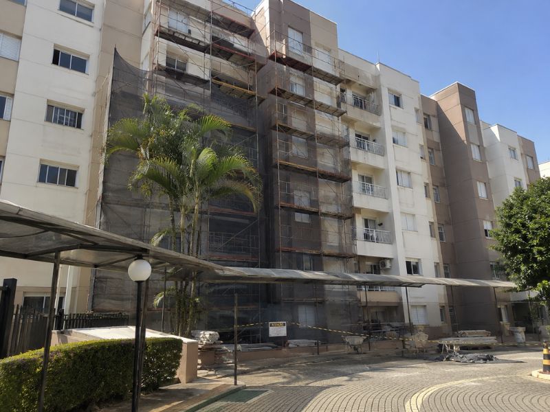 Fachada de prédio residencial em reforma com andaimes e telas de proteção, palmeiras e área verde em frente ao edifício, marquise e área comum de condomínio, céu azul, fotografia realista de reforma predial em condomínio