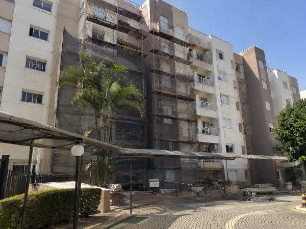 Fachada de condomínio residencial em reforma com andaimes e telas de proteção, palmeiras e área comum ao redor, céu azul, fotografia realista de obra em condomínio