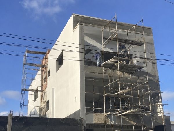Reforma de fachada predial com andaimes metálicos, trabalhadores aplicando revestimento e pintura, prédio em tons claros, céu azul, fotografia realista de obra de reforma