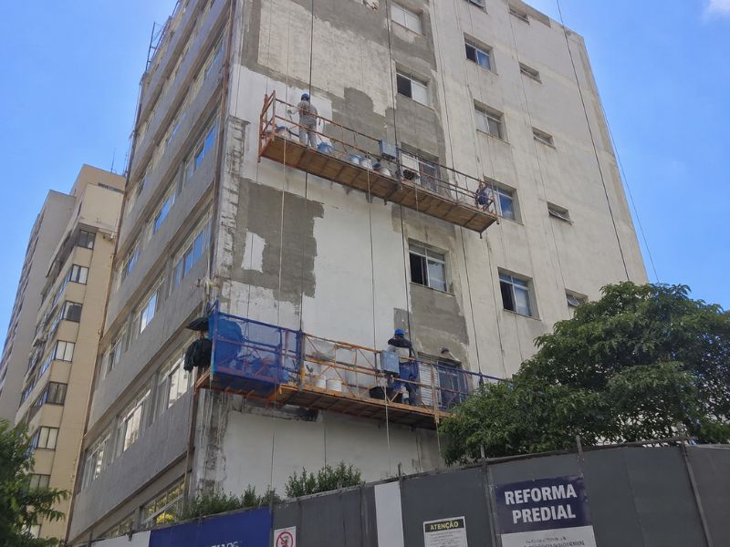 Prédio residencial em reforma de fachada com dois balancins suspensos por cordas e estrutura metálica laranja, trabalhadores com uniforme azul aplicando reboco e revestimento na fachada, tapume cinza na base com placa 'Reforma Predial', céu azul ao fundo, fotografia realista de obra predial em Porto Alegre e Região Metropolitana