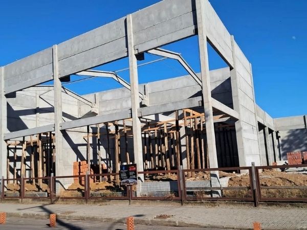 Estrutura de concreto pré-moldado sendo montada em galpão industrial, pilares e vigas de concreto aparente contra céu azul, canteiro de obras com tapume, fotografia realista de construção civil